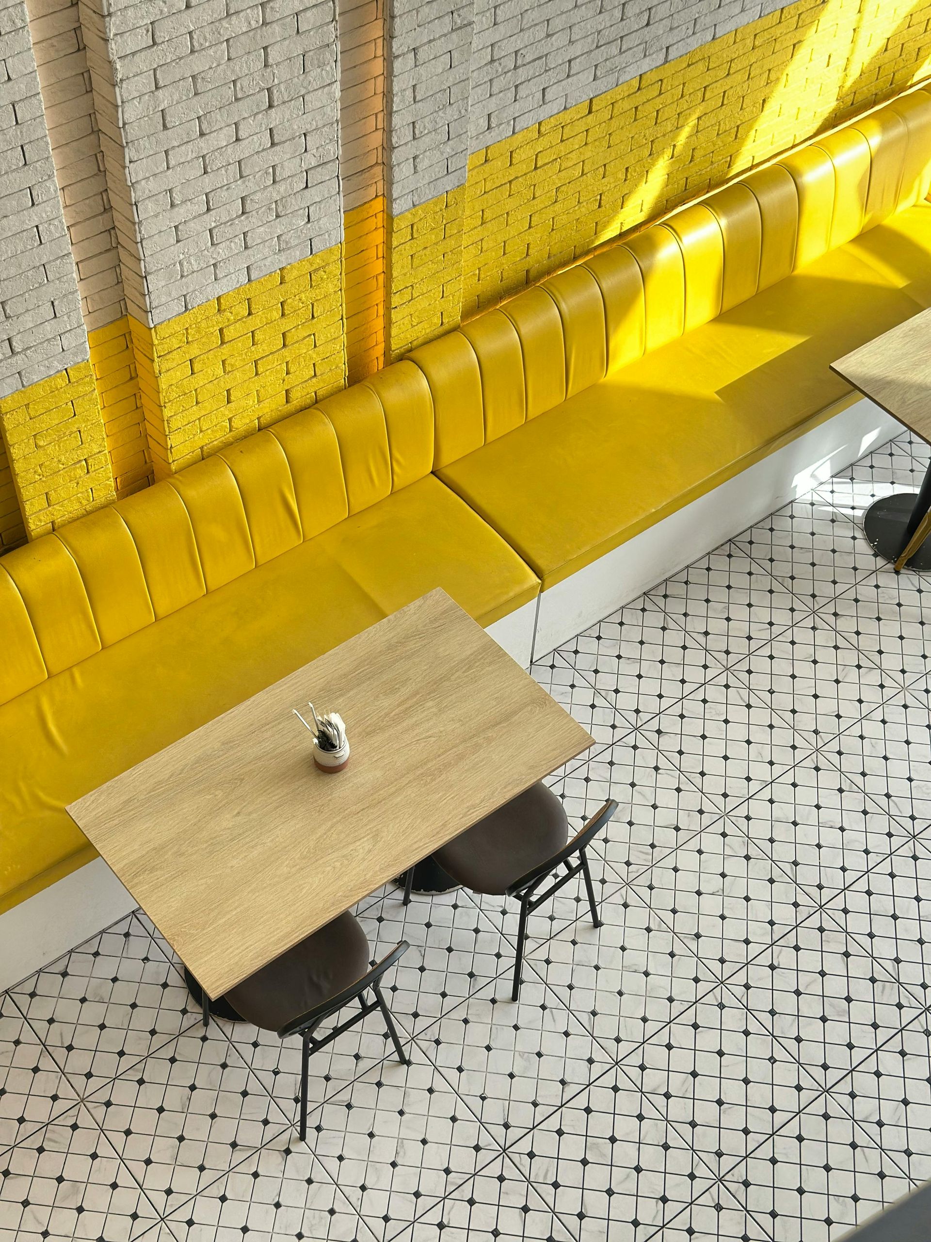 An overhead view of a cafe seating area with a long yellow bench, a light wooden table, and patterned tiled flooring.