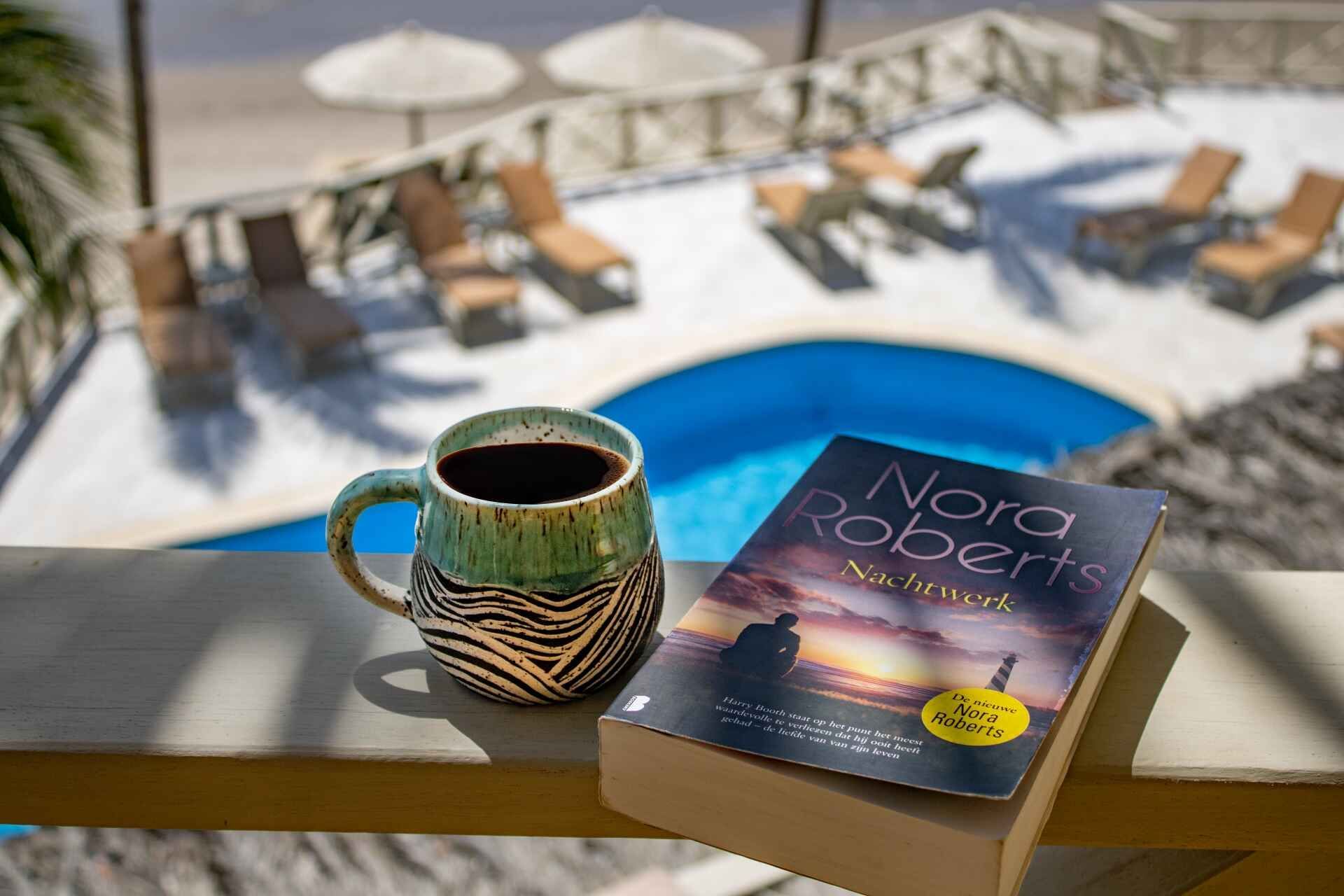 Coffee mug and a book by Nora Roberts on a balcony overlooking a pool and beach.