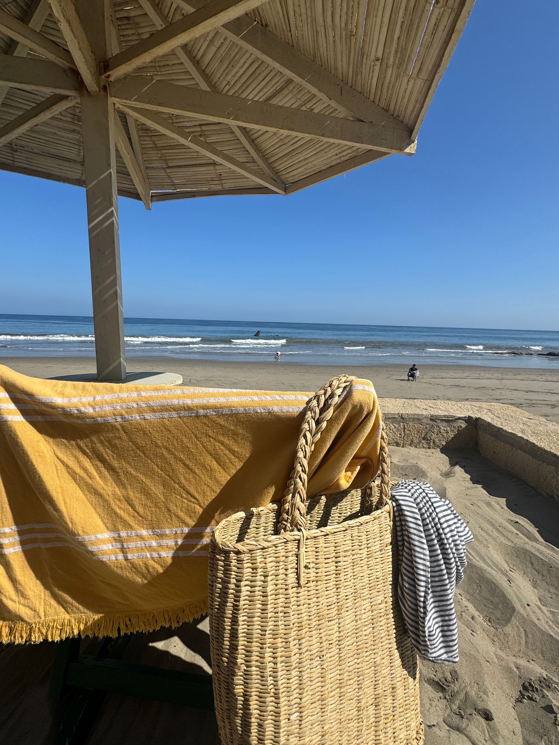 Beach scene: yellow towel, wicker bag, umbrella, ocean, blue sky.