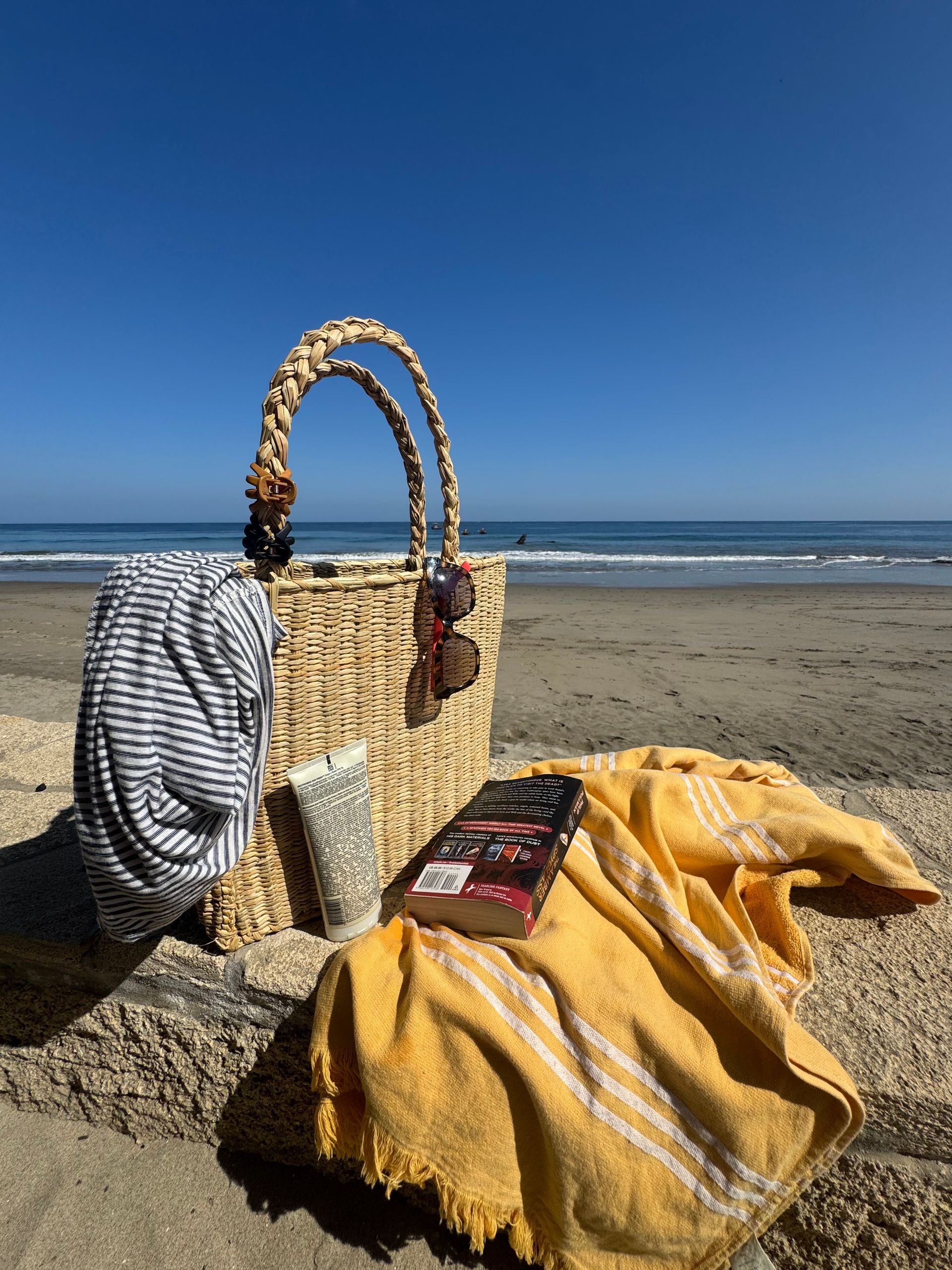 Beach scene: Straw bag, book, and towel on sand, ocean in background under blue sky.