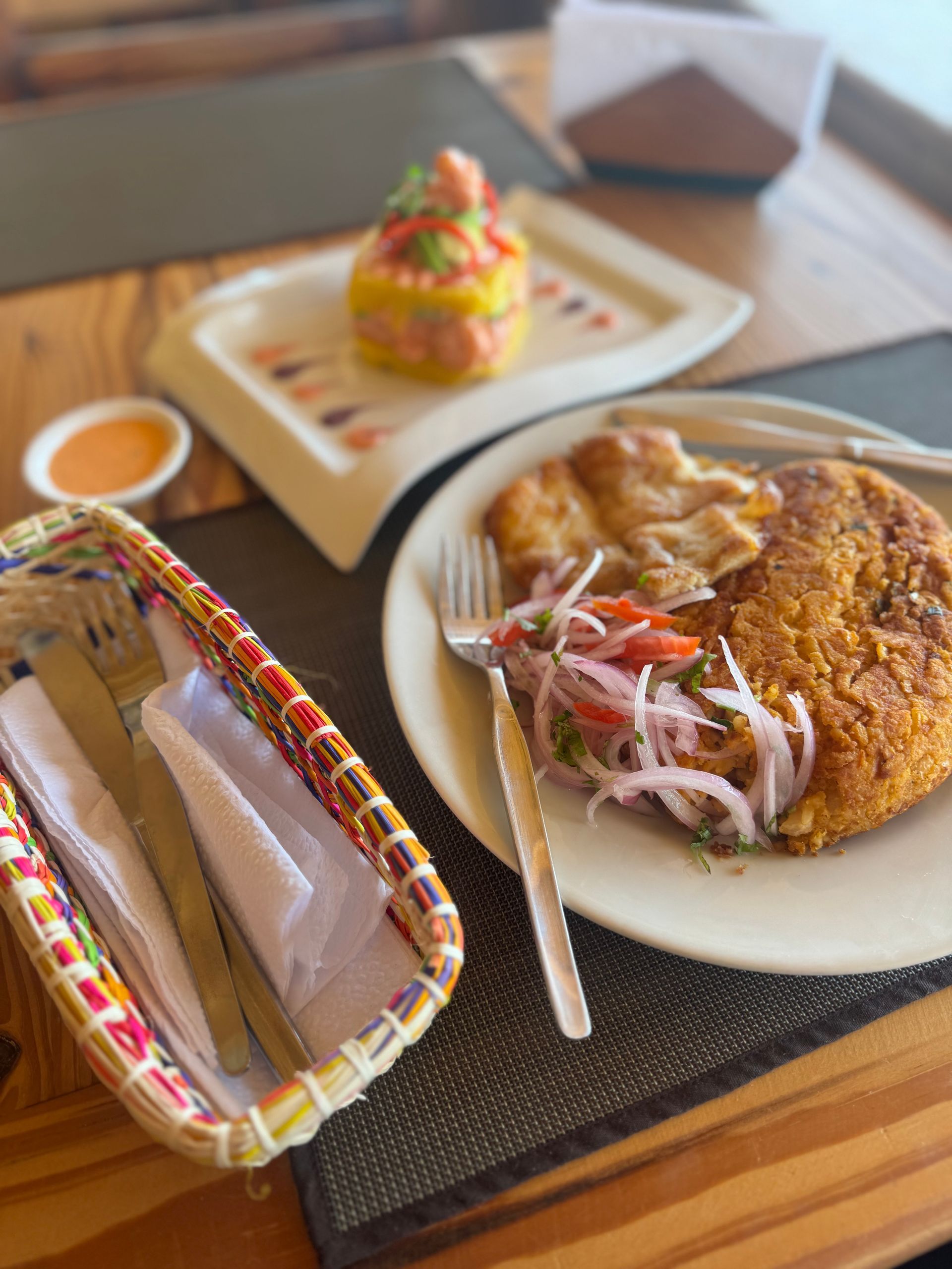 Plate of fried food with salad, ceviche on a plate, cutlery basket, and sauce on a table.