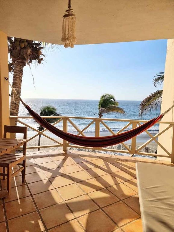Balcony with a hammock overlooking the ocean; palm trees and blue sky in the background.