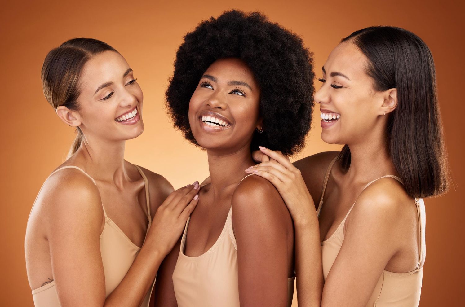Three women smiling, embracing, on a brown background. They wear neutral-toned tops, skin tones vary.