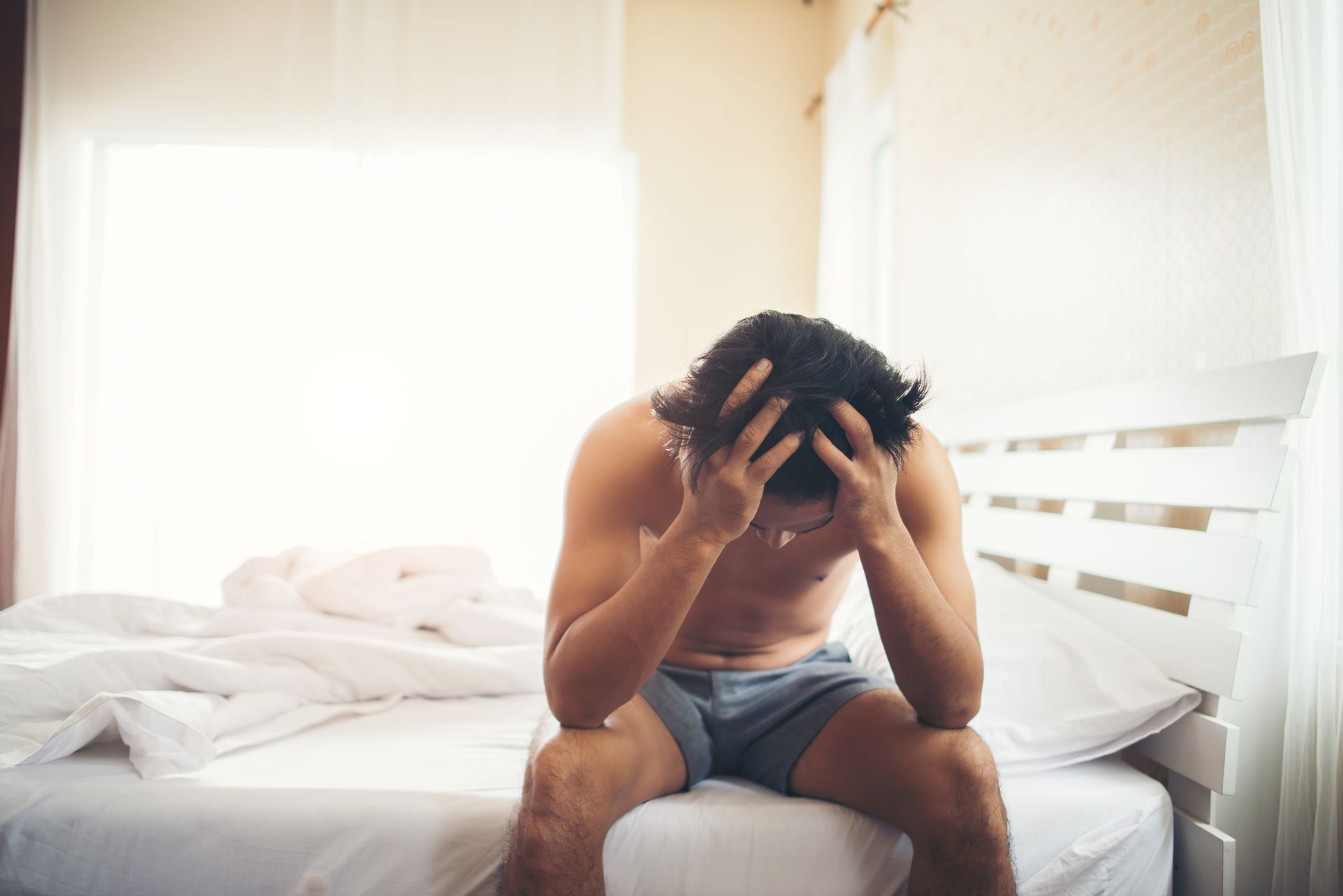 A shirtless man is sitting on a bed with his head in his hands.