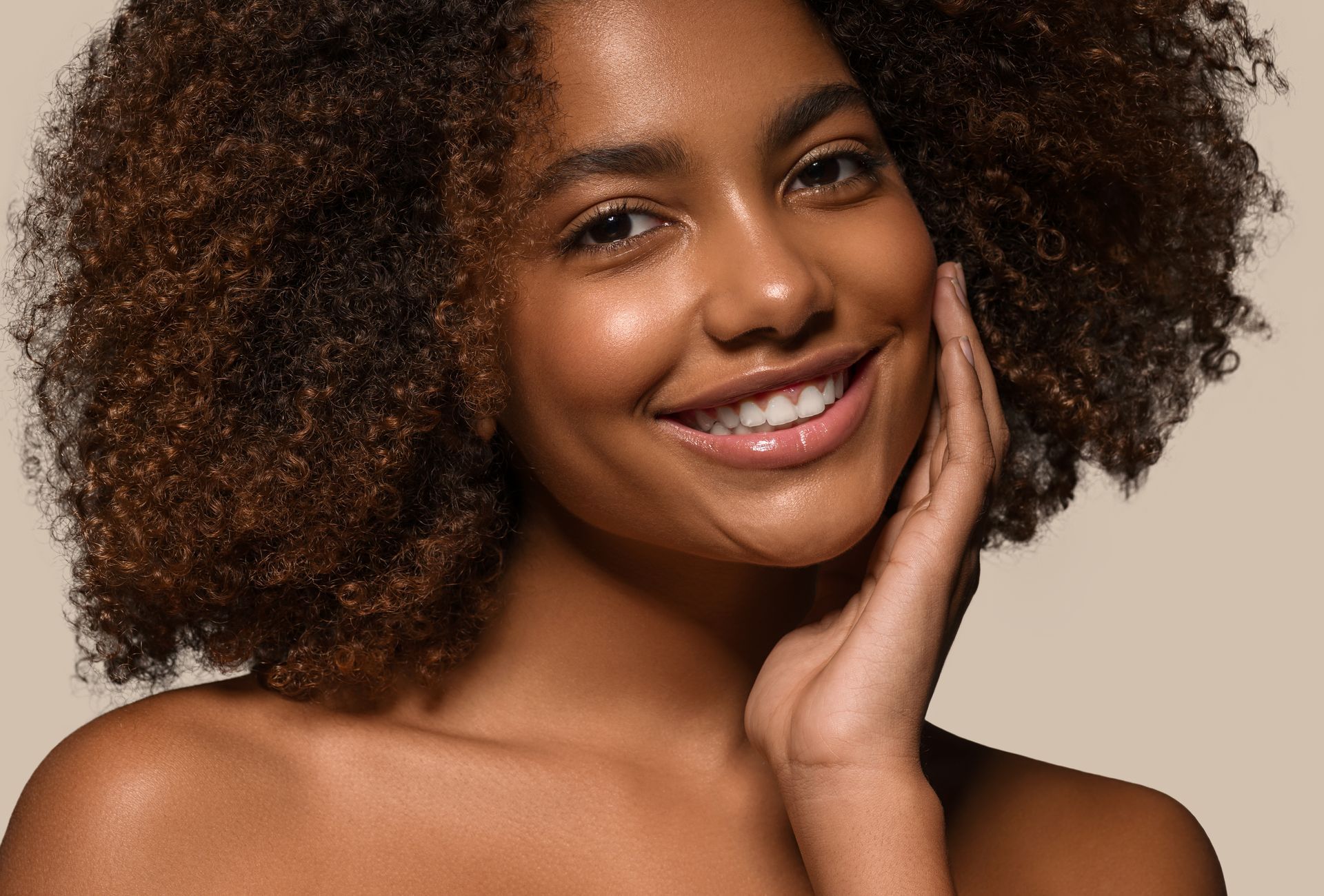 A woman with curly hair is smiling and touching her face.