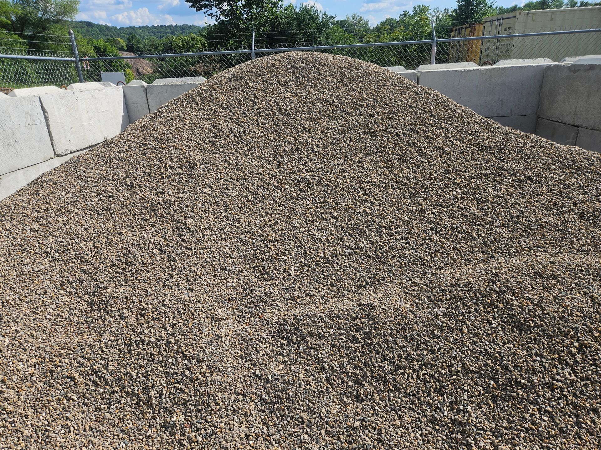 Pile of gray gravel within concrete blocks, outdoors under a bright sky.