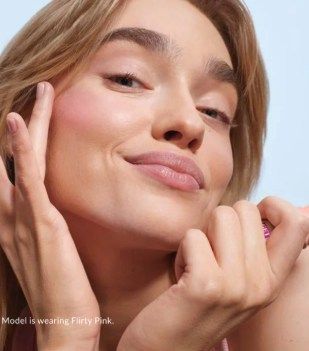 Woman applies blush; pink tones, blue background. Model is wearing