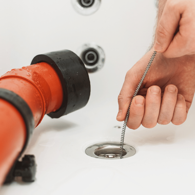 Person unclogging a white bathtub drain with a chain tool, next to a red pipe with a black fitting.