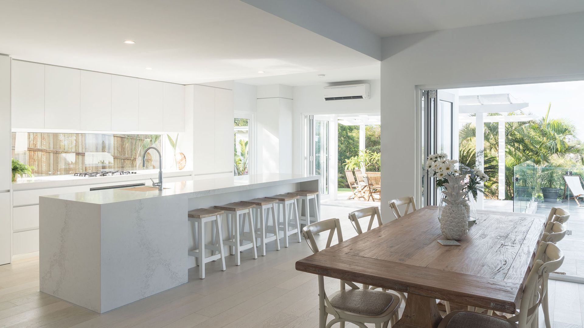 White Modern Kitchen With Island and Bar Stools, Leading to Dining Area With Wooden Table — Akyrah Kitchens in Gympie, QLD