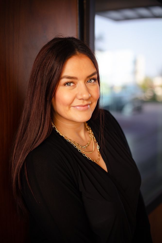A woman in a black dress and gold necklace is standing in front of a window.