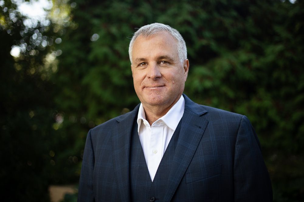 A man in a suit and vest is standing in front of trees.