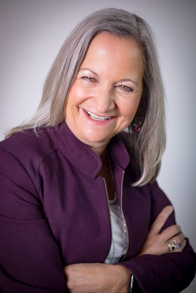 A woman with gray hair is wearing a purple jacket and smiling with her arms crossed.