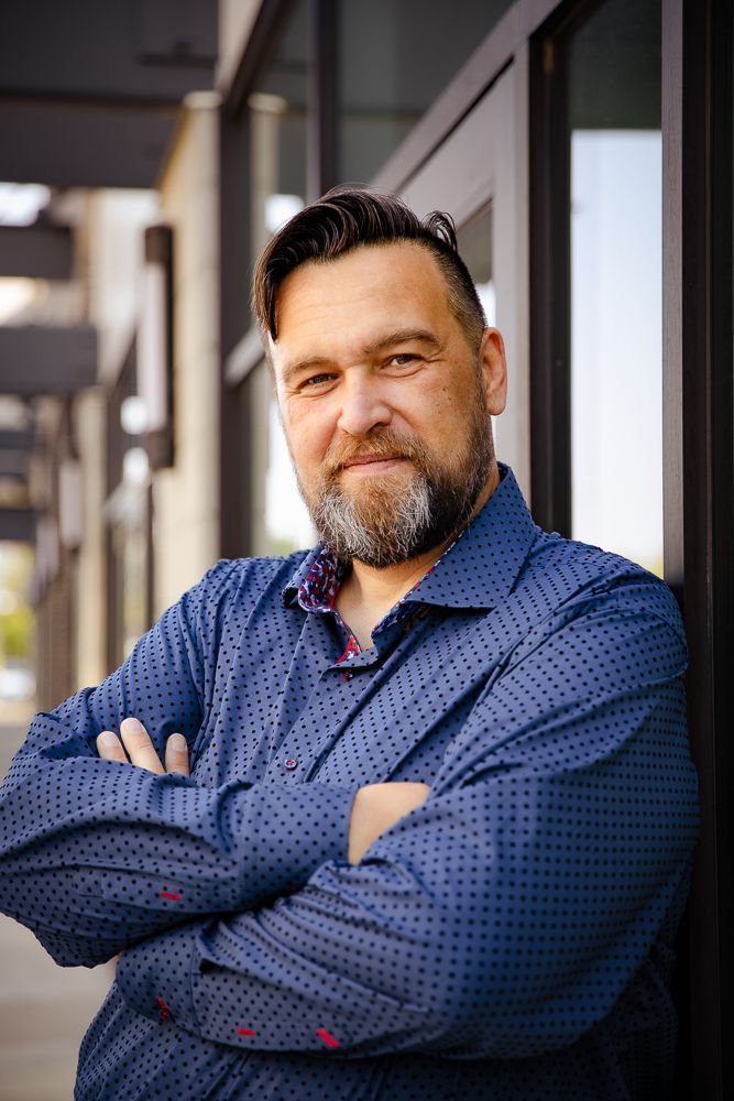 A man with a beard is standing with his arms crossed in front of a building.