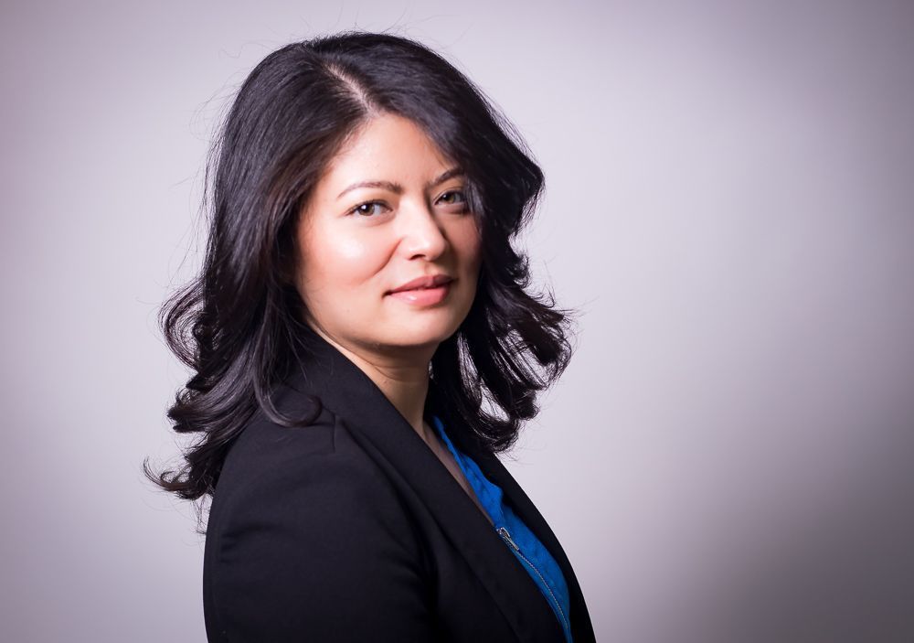 A woman in a black jacket and blue shirt is looking at the camera.