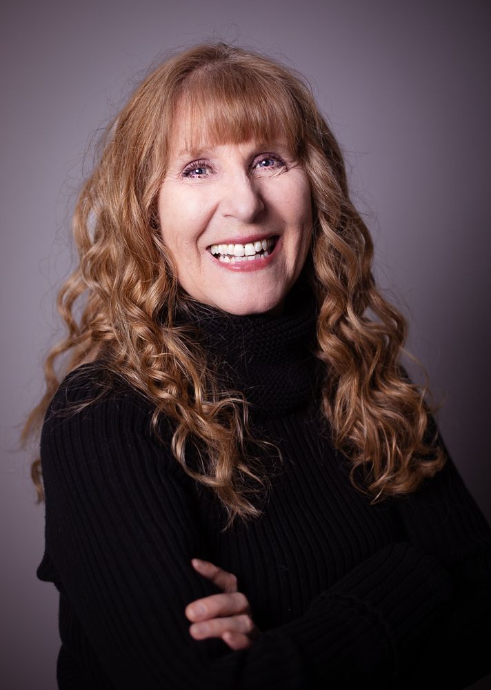 A woman with curly hair is wearing a black turtleneck and smiling.