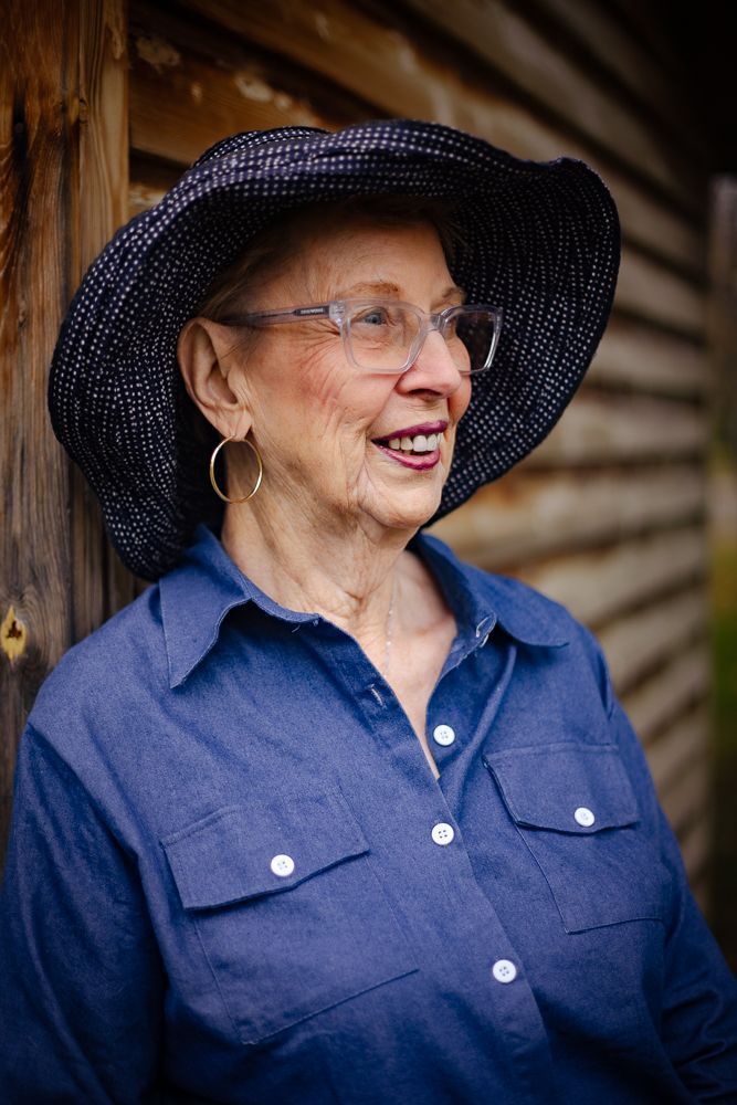 An older woman wearing a hat and glasses is leaning against a wooden wall.