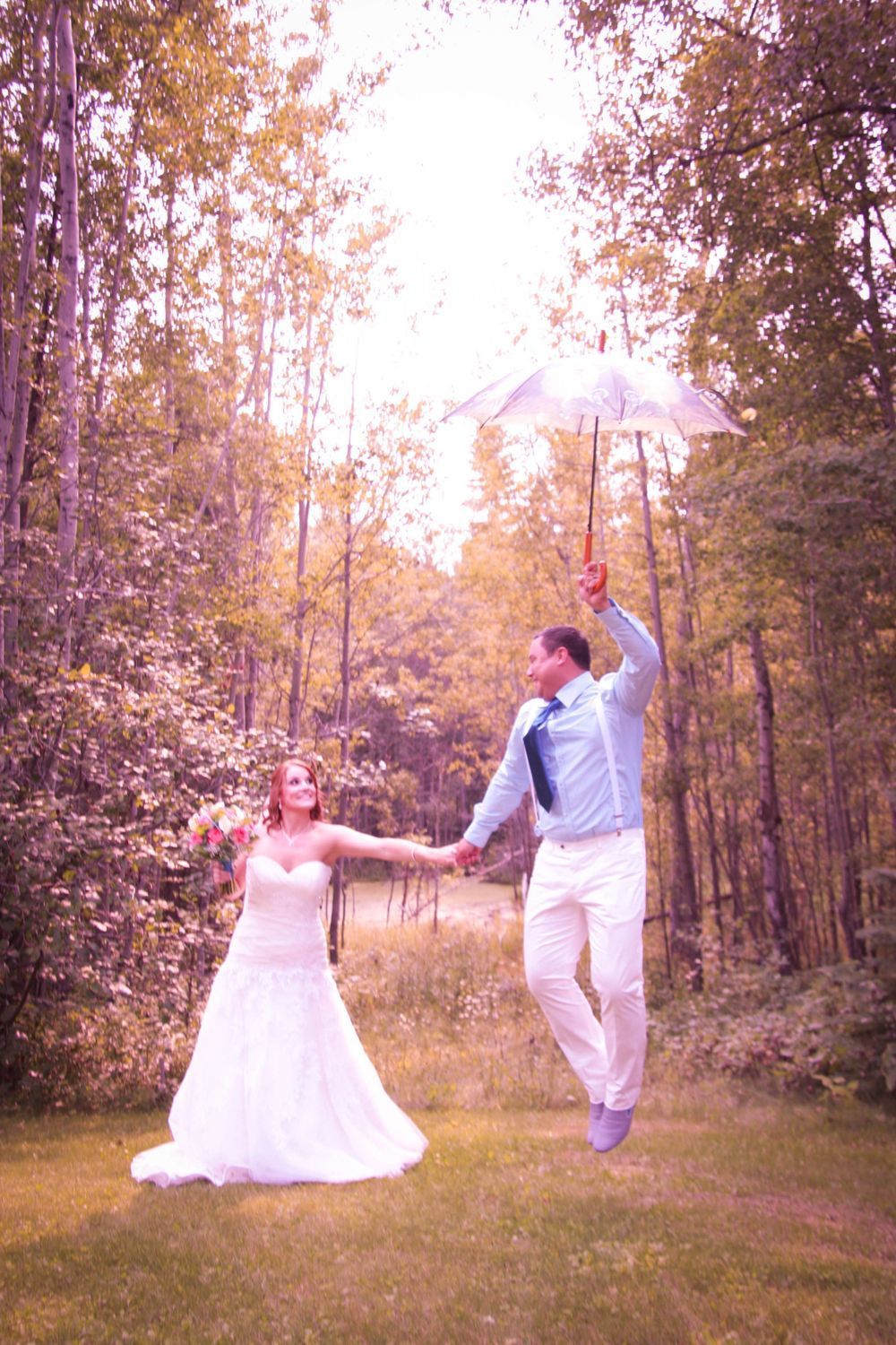 A bride and groom are holding hands and jumping in the air while holding an umbrella.