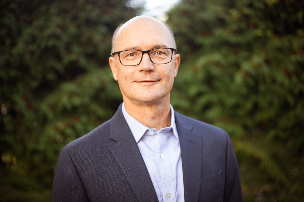A man in a suit and glasses is standing in front of trees.