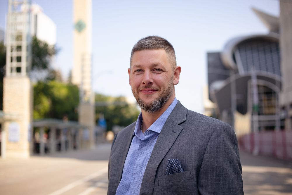 A man in a suit and blue shirt is standing in front of a building.