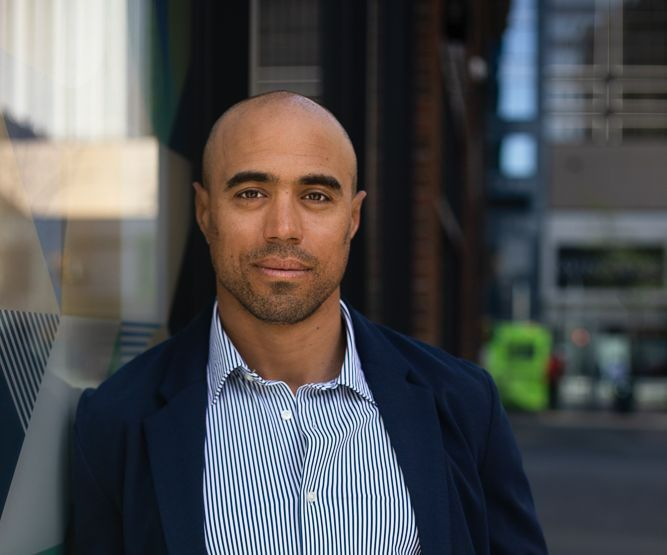 A bald man wearing a blue jacket and striped shirt is standing in front of a building.