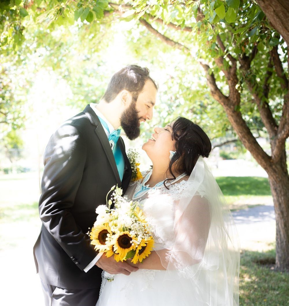 A bride and groom are kissing under a tree while the bride is holding a bouquet of sunflowers.