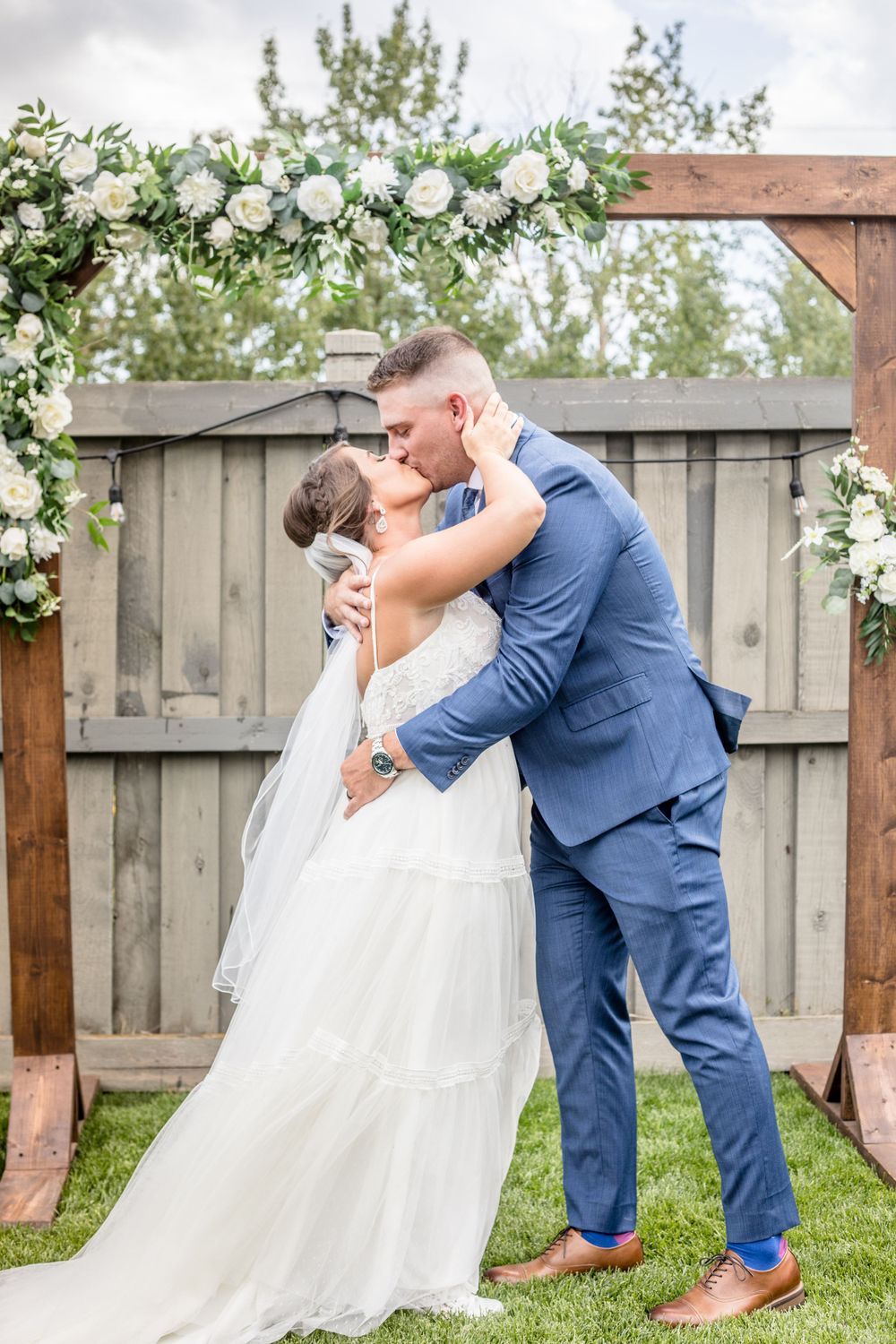 A bride and groom are kissing during their wedding ceremony under a wooden arch.