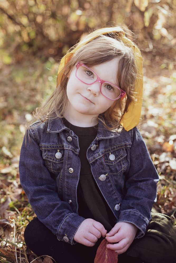 A little girl wearing glasses and a denim jacket is sitting in the leaves.
