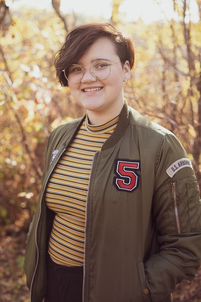 A young woman wearing a bomber jacket and a striped shirt is smiling for the camera.