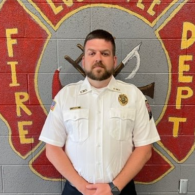 A man in a fire department uniform stands in front of a fire department logo
