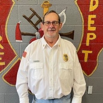 A man in a white shirt and glasses is standing in front of a fire department logo.