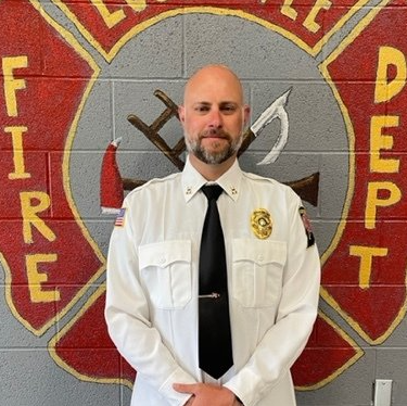 A man in a white shirt and black tie stands in front of a fire department logo