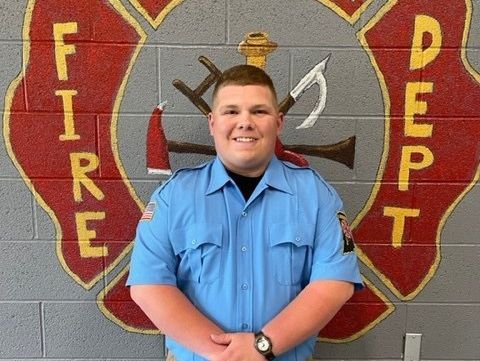 A man in a blue shirt stands in front of a fire department logo