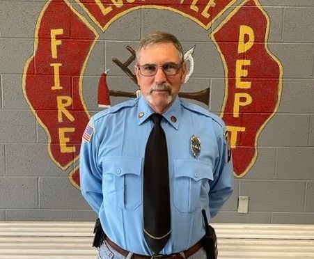A man in a blue shirt and black tie stands in front of a fire department logo