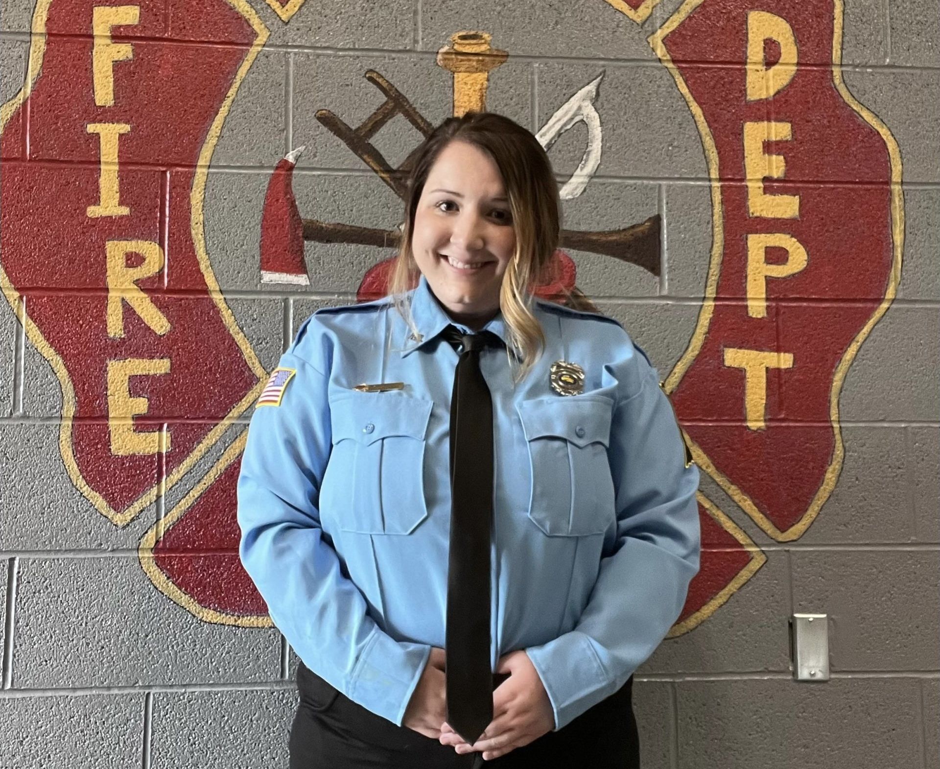 A woman in a blue uniform and tie is standing in front of a fire department logo.