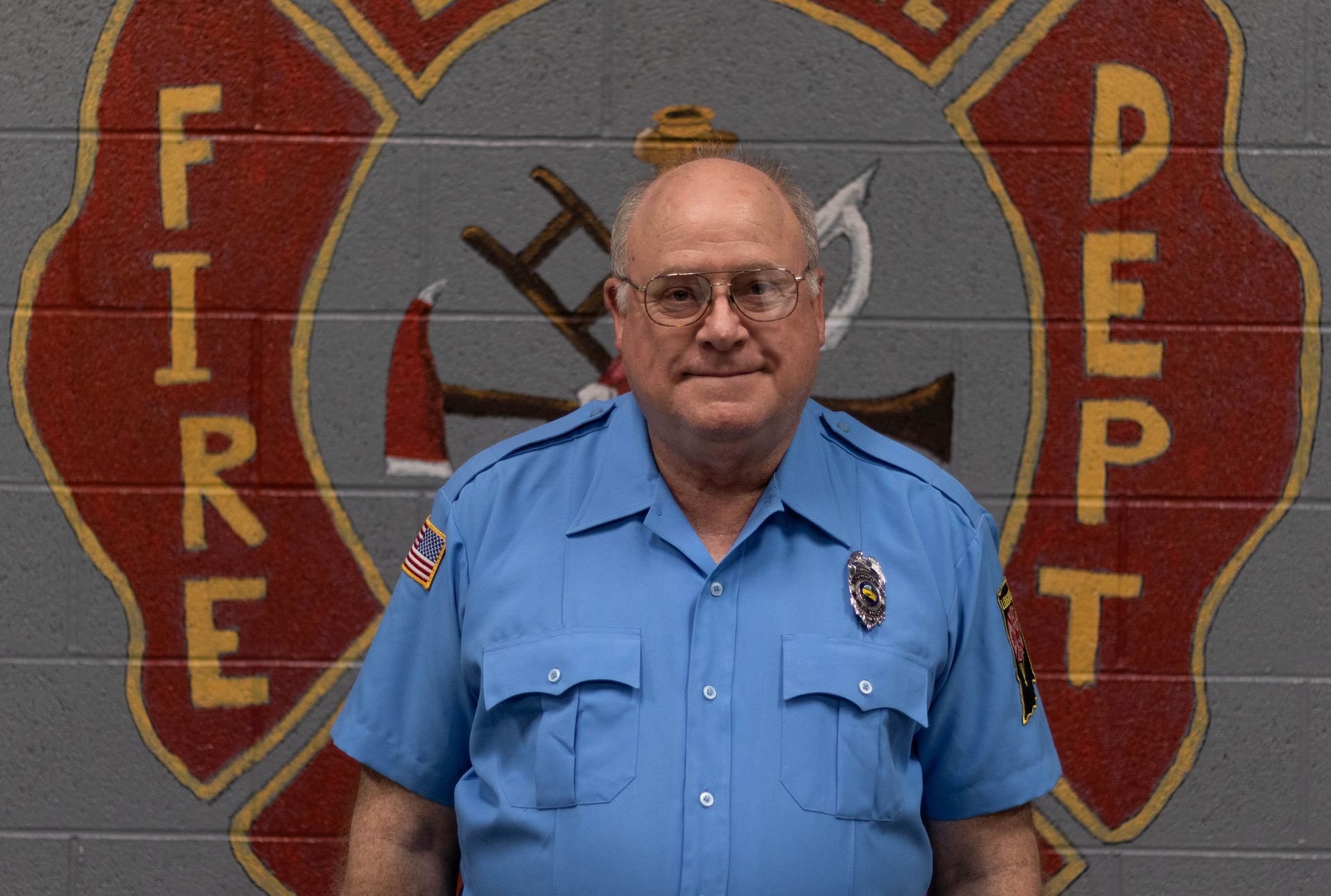 A man in a blue uniform stands in front of a fire department logo