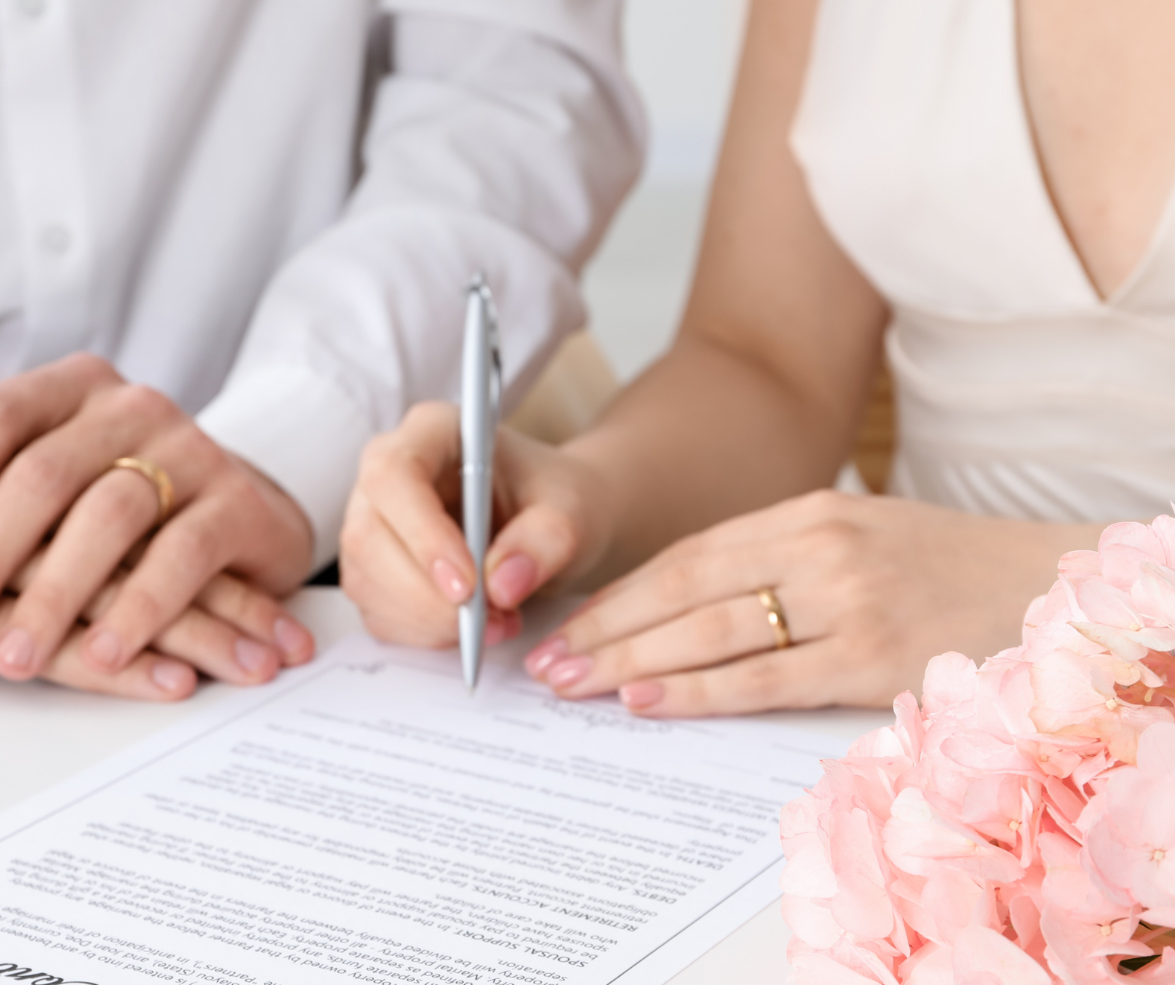 Couple signing marriage documents with rings visible, near a pink floral arrangement.