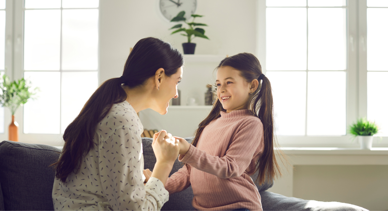 Woman and girl smiling, holding hands on a couch near a window. Sunlight illuminates them.