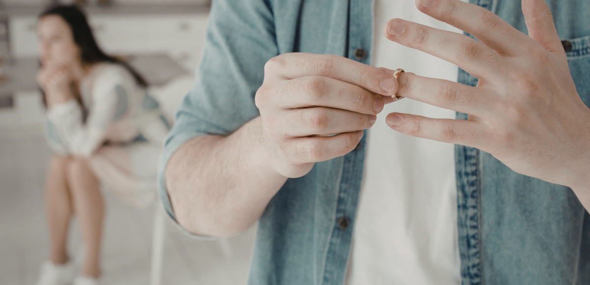 A person removes a ring, while a blurred person sits in the background.