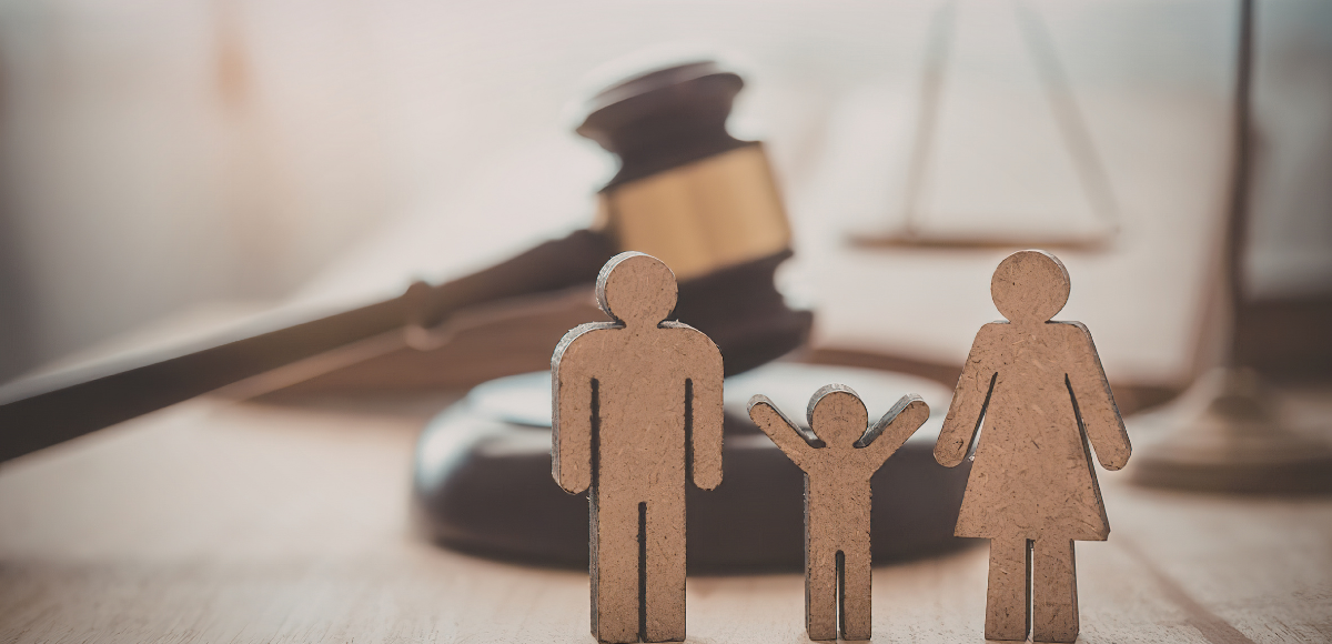 A gavel and scales of justice sit in the background behind small, cardboard cutouts representing a family unit.
