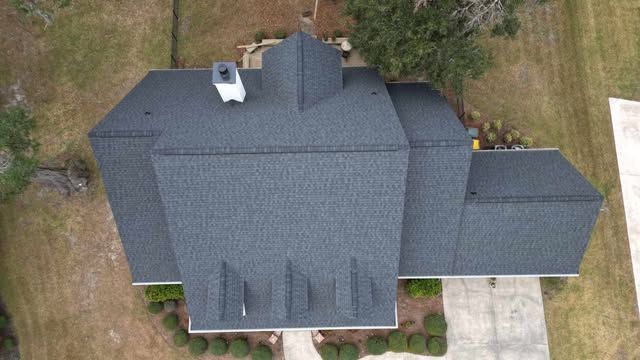 Overhead view of a house with a dark gray shingled roof, a white chimney, and surrounded by green grass.