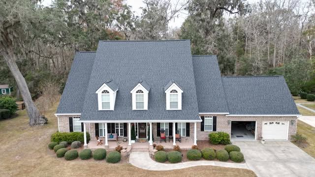 Gray-roofed brick house with three dormers, a porch, and a two-car garage. Landscaping and trees surround.