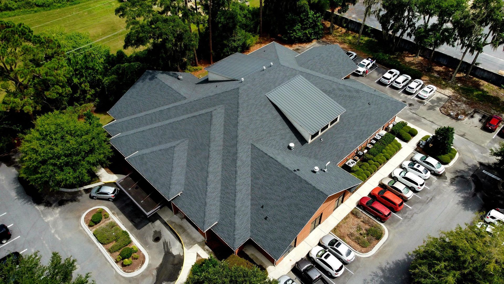 Aerial view of a gray-roofed, one-story building with a parking lot and surrounding greenery.