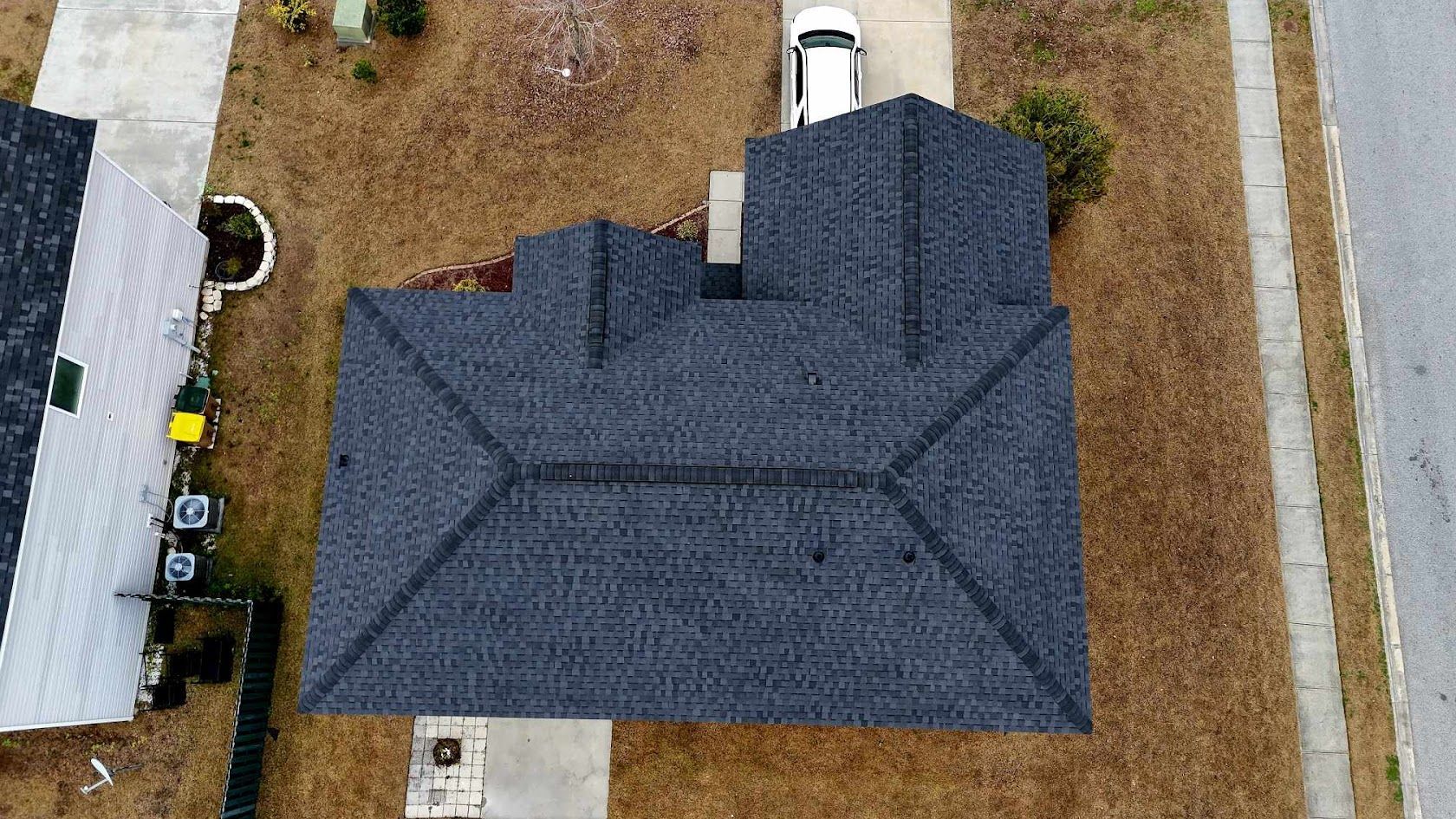 Aerial view of a dark gray shingled house roof with a driveway and car in front, surrounded by brown grass and a sidewalk.
