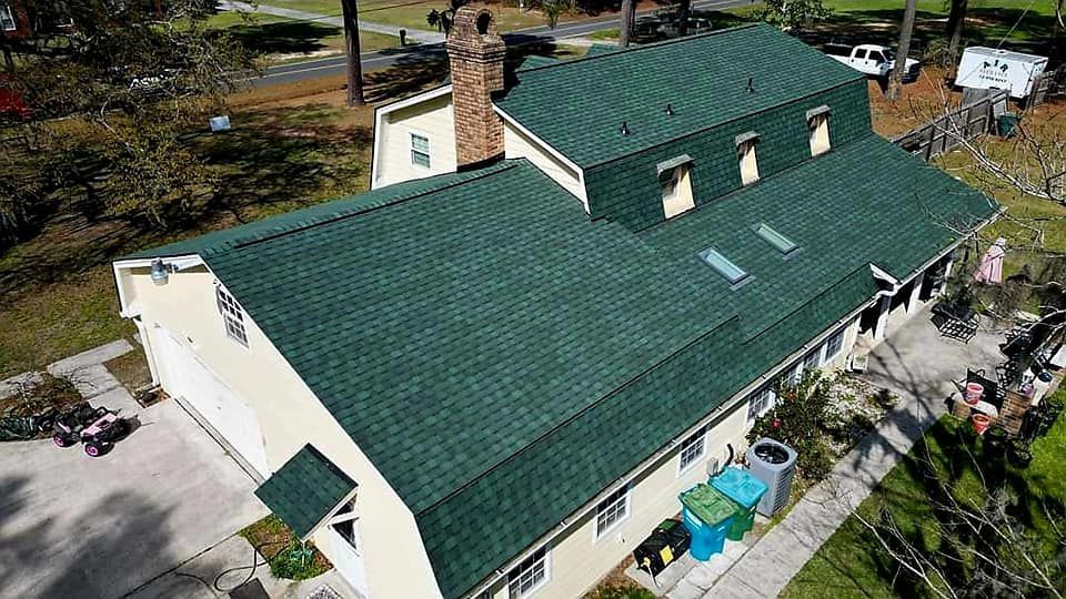 Aerial view of a house with a green roof, off-white exterior, and surrounded by greenery.