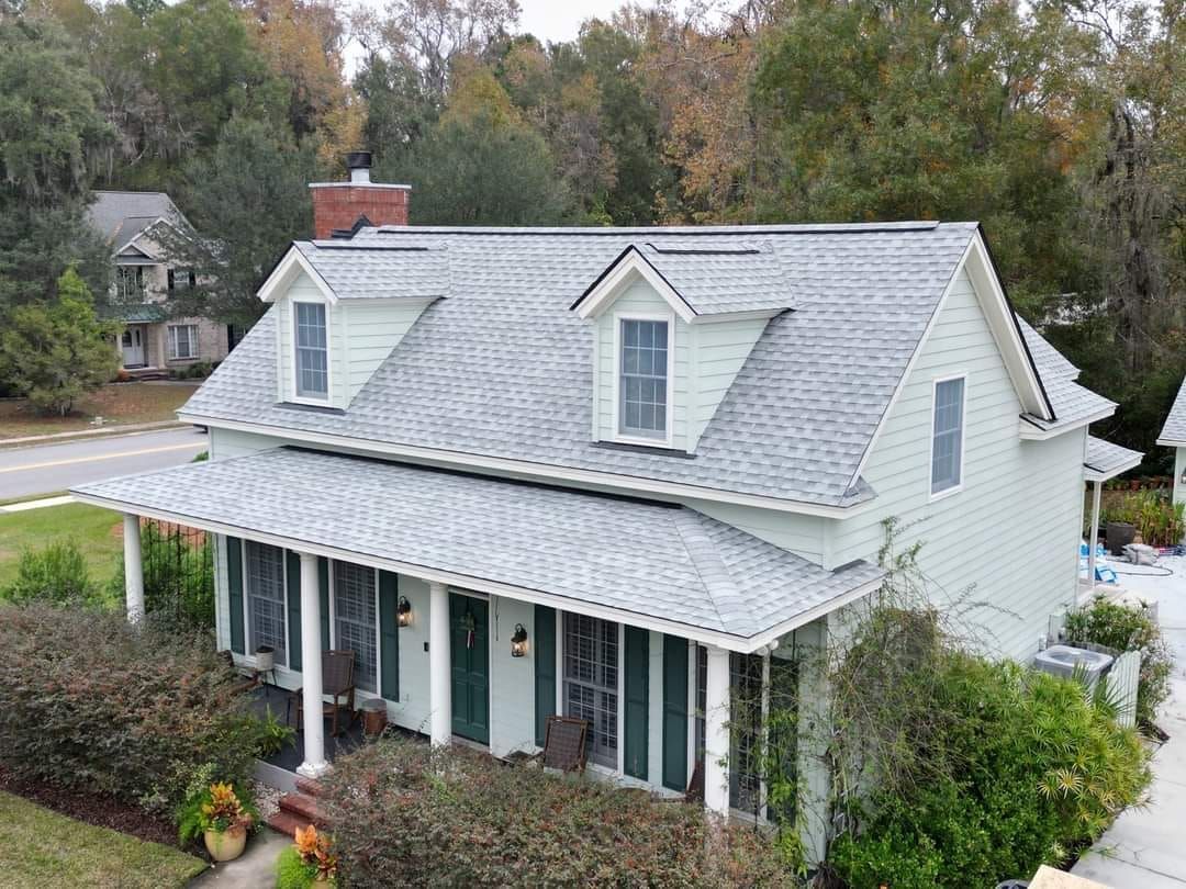 A light green two-story house with a porch and gray roof, two dormers, and surrounding greenery.