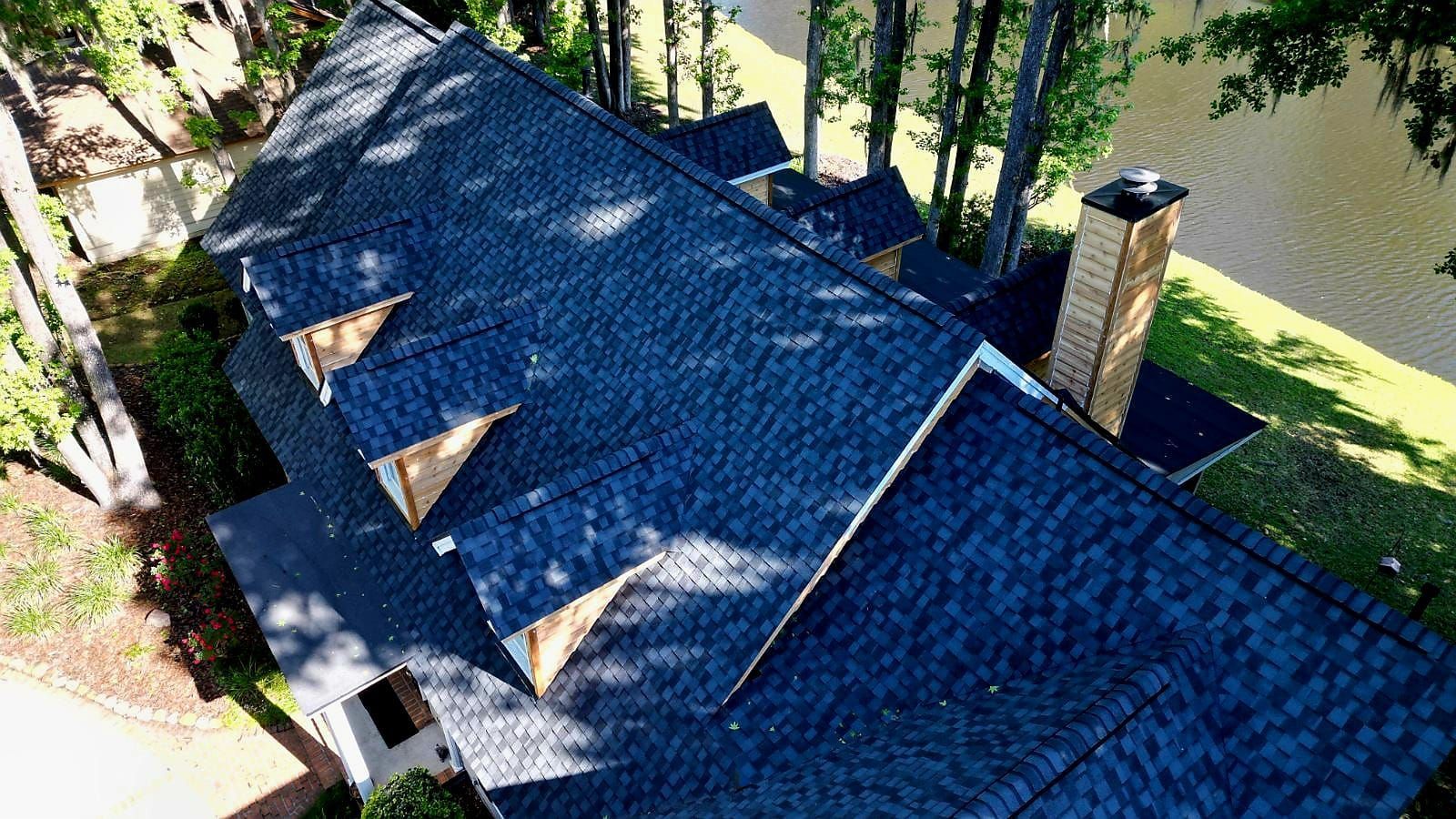 Aerial view of a dark blue shingled roof with a chimney and dormers, near trees and water.