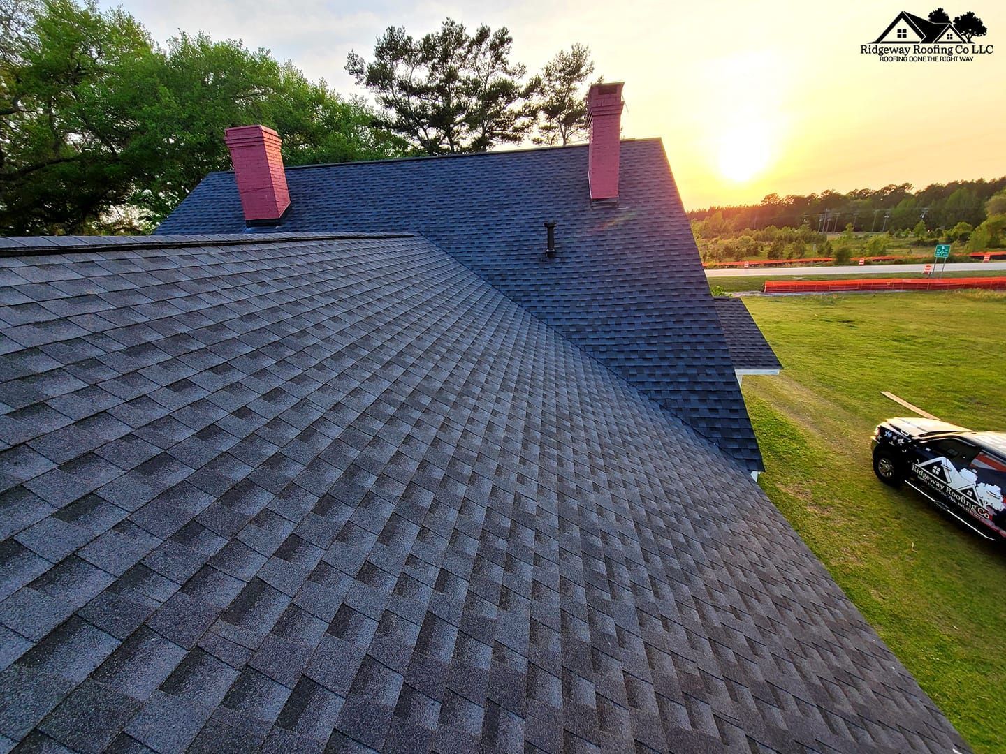 Newly shingled dark gray roof with red chimneys, a car on the grass, and a sunset in the background.
