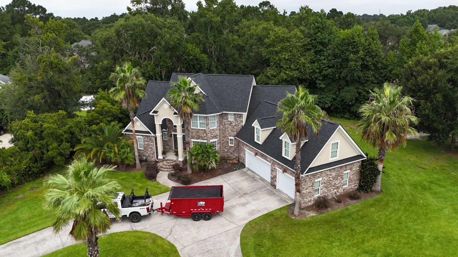 A white truck towing a red trailer in front of a large brick house with a dark roof and palm trees.