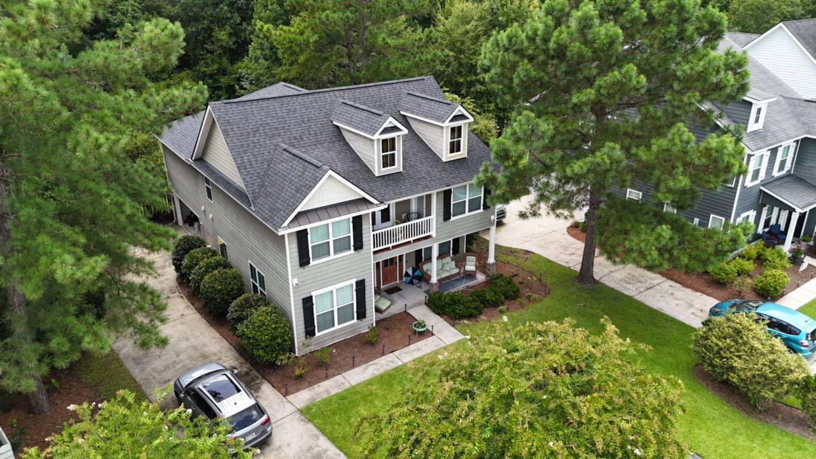 Two-story gray house with black shutters, dormer windows, and a front porch, surrounded by trees and lawn.