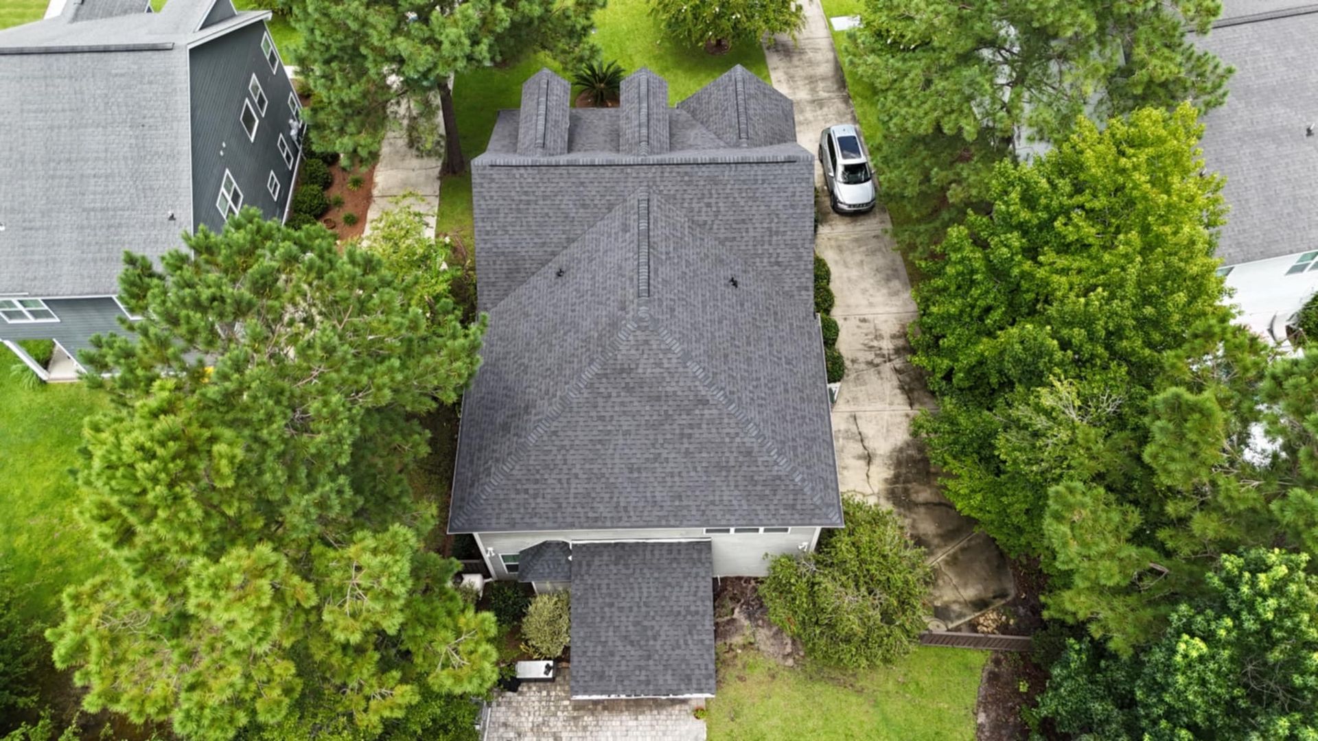 Aerial view of a house with a gray roof surrounded by green trees and a white car in the driveway.